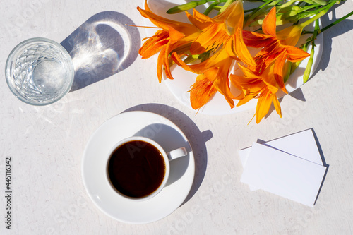Mockup of business cards, a cup of coffee, a glass of water and a bouquet of orange lilies on a white table. Hard long shadows. Flat lay