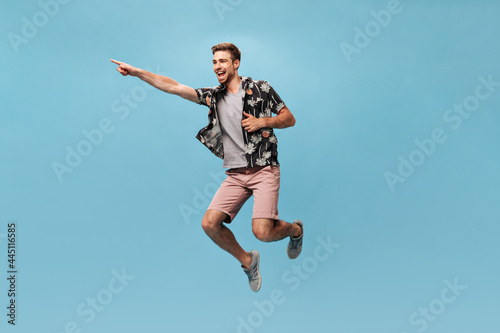 Cool fashionable man with beard in grey t-shirt, printed shirt and pink shorts looking away, rejoices and jumping on blue backdrop..