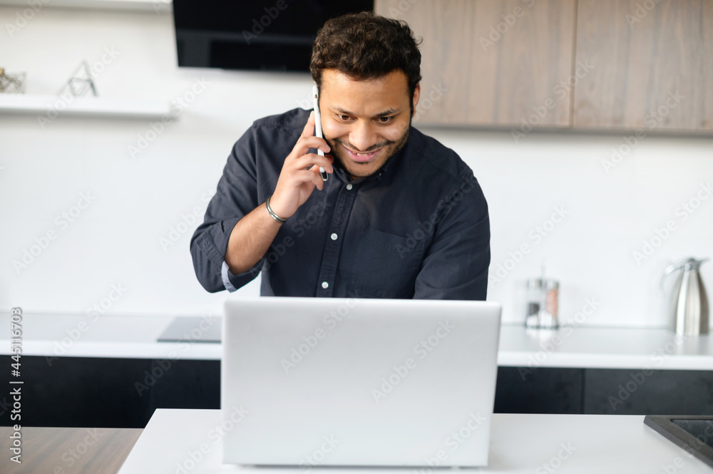 Busy mixed-race young man in smart casual shirt using laptop computer for remote work, a guy is talking on the phone and typing in same time standing in the kitchen at home, work on the distance Busy mixed-race young man in smart casual shirt using laptop computer for remote work, a guy is talking on the phone and typing in same time standing in the kitchen at home, work on the distance