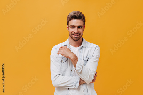Fashionable bearded guy with stylish hairstyle in white modern jacket smiling and looking into camera on isolated backdrop..