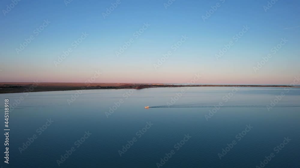 A white boat sails on Lake Balkhash at sunset. A beautiful long train on the water from the motor. The sky gradient is from orange to blue. Smooth water. Travel on a speed bot. Bertys Bay. Kazakhstan A white boat sails on Lake Balkhash at sunset. A beautiful long train on the water from the motor. The sky gradient is from orange to blue. Smooth water. Travel on a speed bot. Bertys Bay. Kazakhstan