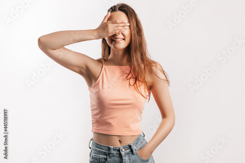Young beautiful girl smiles and covers of her face with her hand palm on a white isolated background. Positive brunette woman in orange top