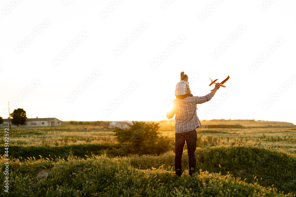 Family single father with his daughter at sunset in a field on a summer evening launching a toy airplane into the air. baby girl sitting on dad's shoulders looks the sky.childhood concept