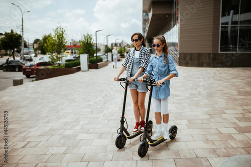 Portrait of a modern young mom and daughter on a day off, having fun riding electric scooters around the city. #445126753