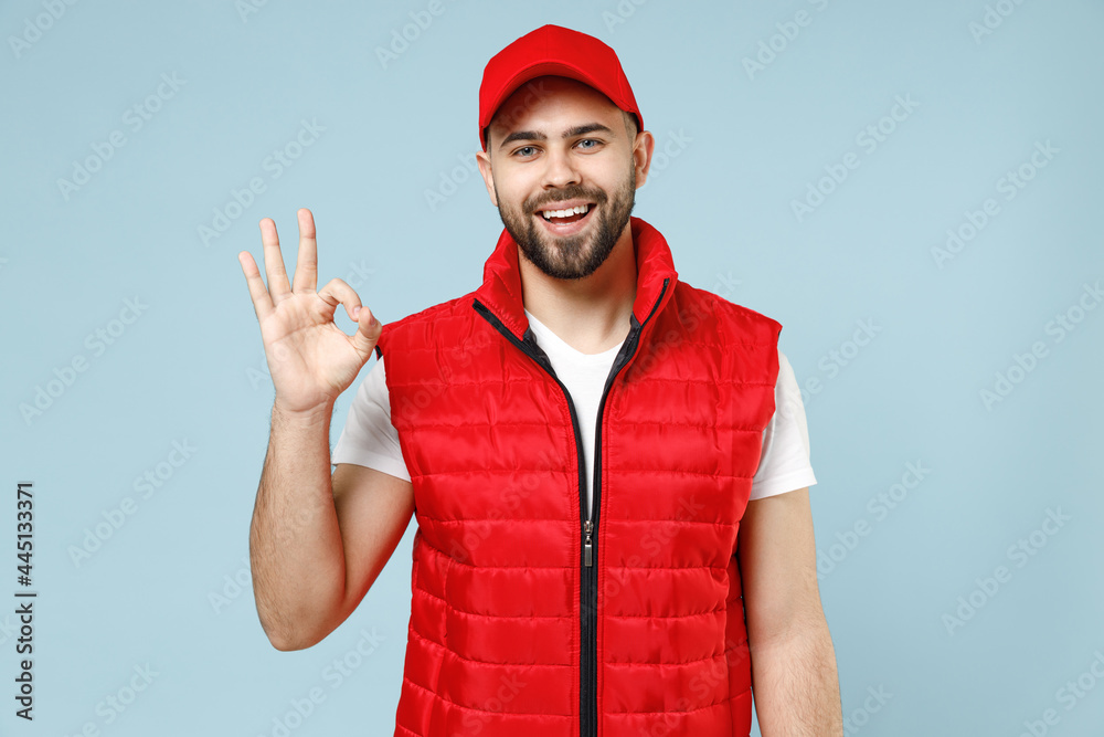 Naklejka premium Professional happy delivery guy employee man in red cap white T-shirt vest uniform workwear work as dealer courier show ok okay gesture isolated on pastel blue color background studio Service concept