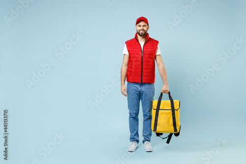 Full size body length delivery guy employee man in red cap white T-shirt uniform work as courier hold yellow thermal food bag backpack isolated on pastel blue color background studio. Service concept. photo