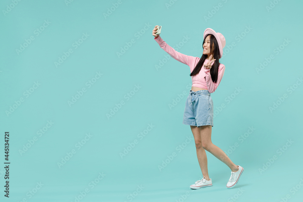 Full size body length fun young brunette asian woman 20s wears pink clothes doing selfie shot on mobile phone showing victory sign strolling isolated on pastel blue color background studio portrait