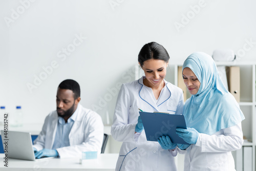 Interracial scientists smiling while holding clipboard near blurred african american colleague with laptop in lab © LIGHTFIELD STUDIOS
