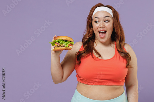 Young fun happy excited fun chubby overweight plus size big fat fit woman in red top warm up training hold fast food eating burger isolated on purple background gym Workout sport motivation concept.
