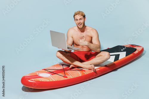 Full length young fun man in red shorts swimsuit sit on sup board use laprop pc computer work online show thumb up gesture isolated on pastel blue background Summer vacation sea rest sun tan concept © ViDi Studio