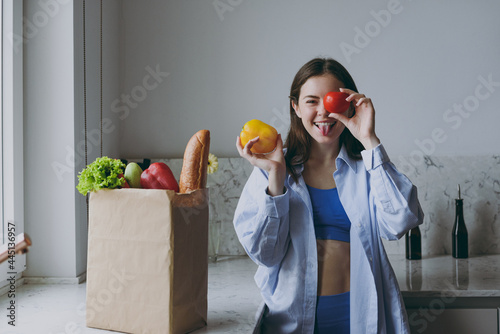 Young fun vegeterian housewife woman 20s in casual clothes blue shirt cover eye with tomato bell pepper papric show tongue cooking food in light kitchen at home alone Healthy diet lifestyle concept photo