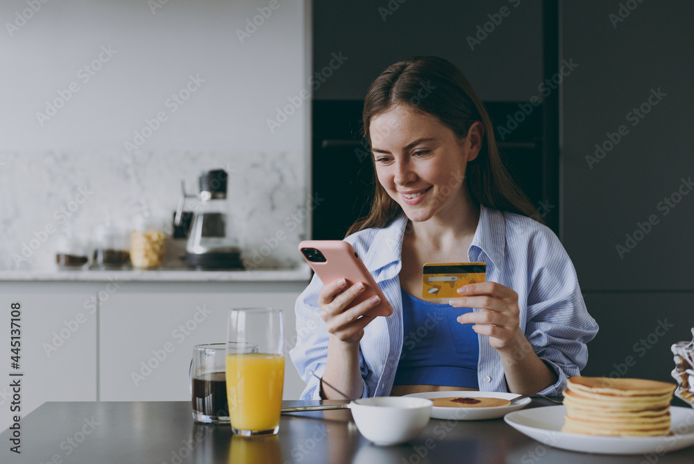 Young housewife woman in casual clothes eat breakfast pancakes using mobile cell phone credit bank card shop online order delivery cook food in light kitchen at home. Healthy diet lifestyle concept Young housewife woman in casual clothes eat breakfast pancakes using mobile cell phone credit bank card shop online order delivery cook food in light kitchen at home. Healthy diet lifestyle concept