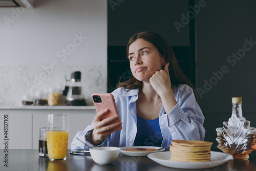 Young pensive housewife woman 20s in casual clothes blue shirt eating breakfast pancakes use mobile cell phone look for recipe cook food in light kitchen at home alone Healthy diet lifestyle concept.