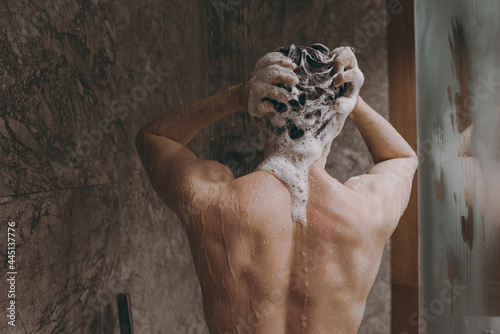 Back rear view young brunet half naked torso topless brunette man 20s taking hot shower wash hair with shampoo indoors in bathroom at home. People healthcare daily morning routine lifestyle concept