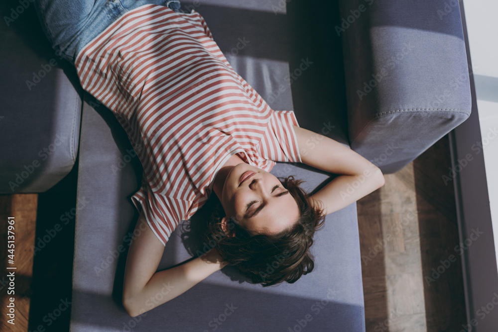 Fototapeta premium Top view from above young woman in casual clothes lying on grey sofa hold hands behind head neck relax take nap during day break on weekends rest indoors flat at home People lifestyle leisure concept