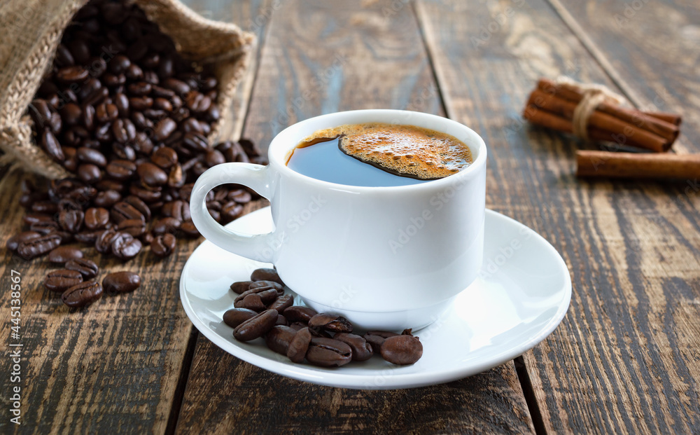 Fototapeta premium Black coffee in a white cup, coffee beans, cinnamon sticks on an old wooden background.
