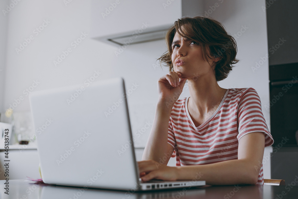 Young puzzled pensive programmer freelancer housewife woman in casual clothes striped t-shirt use laptop pc computer work online prop up chin in light kitchen at home alone People lifestyle concept. Young puzzled pensive programmer freelancer housewife woman in casual clothes striped t-shirt use laptop pc computer work online prop up chin in light kitchen at home alone People lifestyle concept.