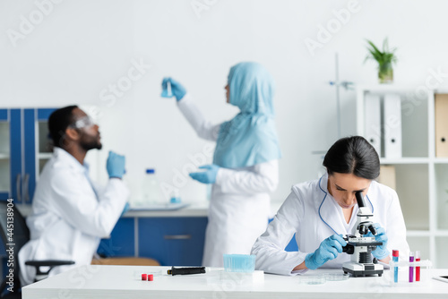 Scientist looking through microscope near blurred interracial colleagues in lab