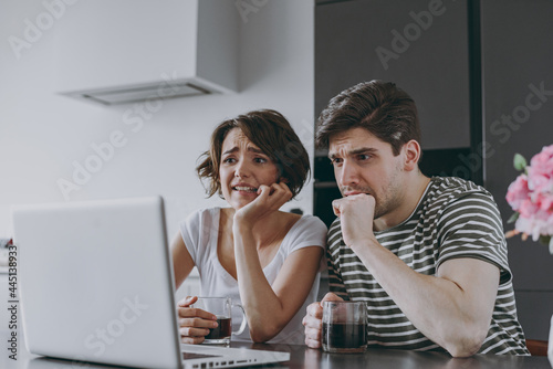 Young sad confused couple two woman man in casual t-shirt clothes sit table drink coffee using laptop pc computer ith virus do oops gesture in light kitchen at home together People lifestyle concept.