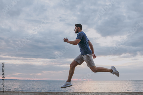 Side view young strong sporty athletic toned fit sportsman man 20s in sports clothes warm up training run fast at sunrise sun dawn over sea beach outdoor on pier seaside in summer day cloudy morning.