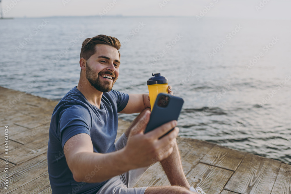 Young sporty strong sportsman man 20s in sports clothes shorts train do selfie shot photo on mobile cell phone drink water at sunrise sun over sea beach outdoor on pier seaside in summer day morning. Young sporty strong sportsman man 20s in sports clothes shorts train do selfie shot photo on mobile cell phone drink water at sunrise sun over sea beach outdoor on pier seaside in summer day morning.