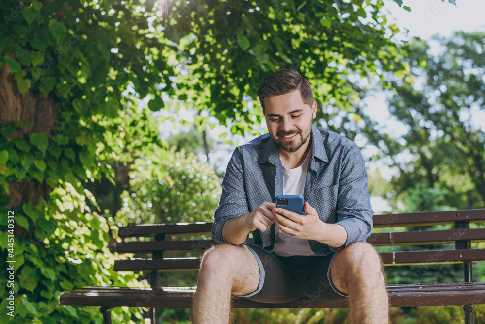 Bottom view smiling satisfied young man in blue shirt sit on bench use mobile cell phone chat online rest relax in spring green city sunshine park outdoors on nature Urban lifestyle leisure concept Bottom view smiling satisfied young man in blue shirt sit on bench use mobile cell phone chat online rest relax in spring green city sunshine park outdoors on nature Urban lifestyle leisure concept
