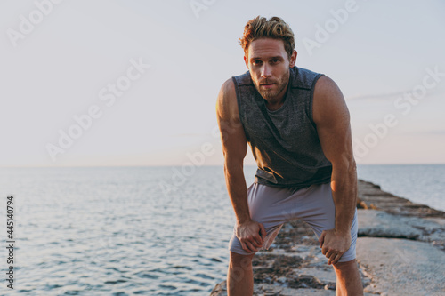 Tired exhausted young strong sporty athletic toned fit sportsman man 20s in sports clothes warm up train look far away at sunrise sun dawn over sea beach outdoor on pier seaside in summer day morning.
