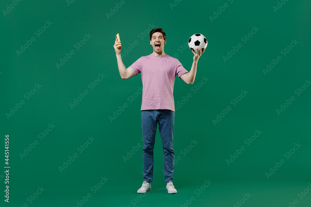 Full size body length fun young man fan in pink t-shirt cheer up football sport team hold hand soccer ball mobile cell phone watch tv live stream scream isolated on dark green color background studio.