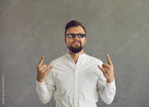 Self assured man wearing funny sunglasses. Studio shot of smirking smug looking student or businessman in white shirt and thug life glasses doing rock sign gesture standing isolated on grey background photo