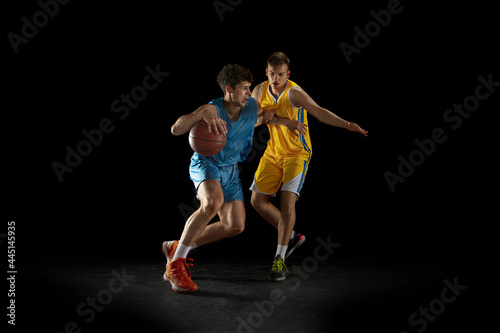 Two basketball players in action and motion isolated on dark black studio background. Advertising concept. Strong Caucasian athletes practicing with basketball ball.