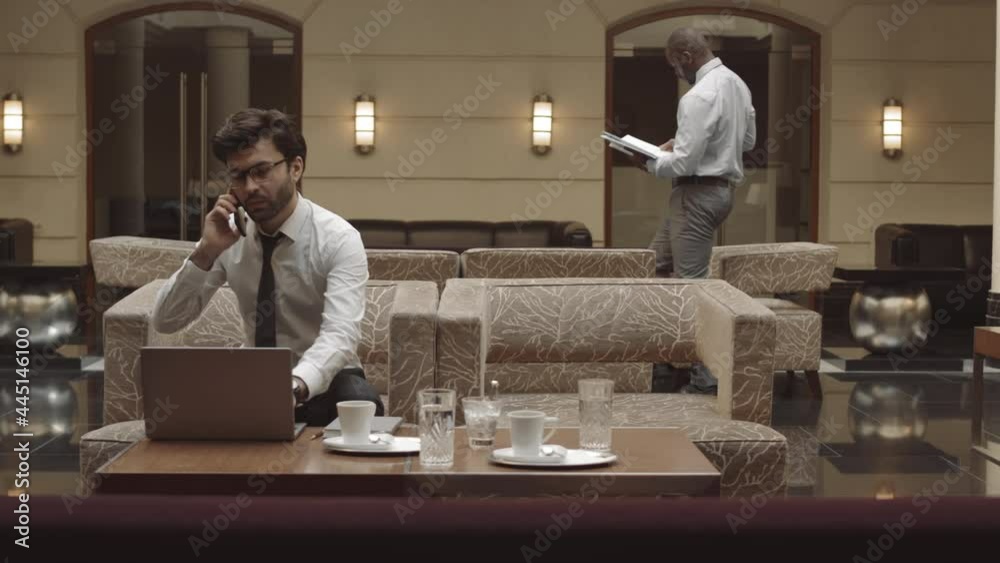 Medium long of young successful CEO sitting in armchair in restaurant with laptop computer on table, talking on mobile phone, his coworker walking on background Medium long of young successful CEO sitting in armchair in restaurant with laptop computer on table, talking on mobile phone, his coworker walking on background