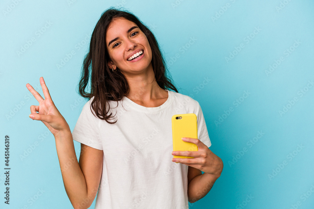 Young caucasian woman holding a mobile phone isolated on blue background joyful and carefree showing a peace symbol with fingers. Young caucasian woman holding a mobile phone isolated on blue background joyful and carefree showing a peace symbol with fingers.