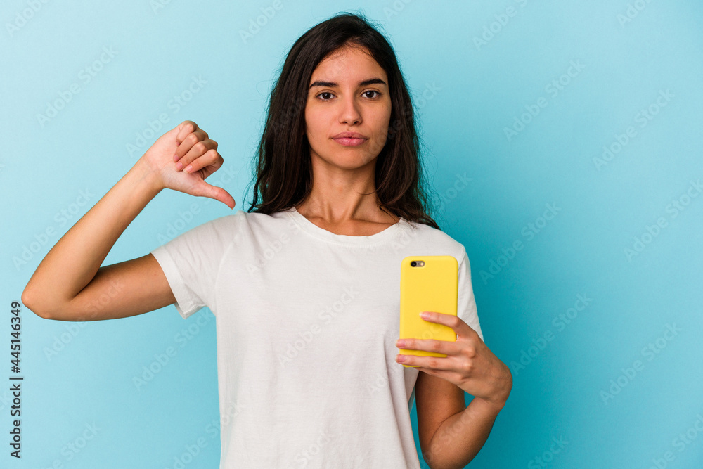 Young caucasian woman holding a mobile phone isolated on blue background showing a dislike gesture, thumbs down. Disagreement concept. Young caucasian woman holding a mobile phone isolated on blue background showing a dislike gesture, thumbs down. Disagreement concept.