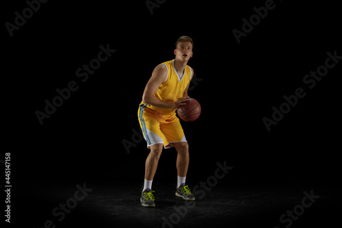 One young man, basketball player with a ball training isolated on dark black studio background. Advertising concept. Fit Caucasian athlete practicing with ball. © master1305