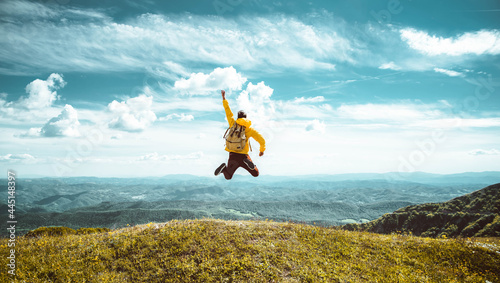 Fotografia Hiker with backpack raising hands jumping on the top of a mountain - Successful