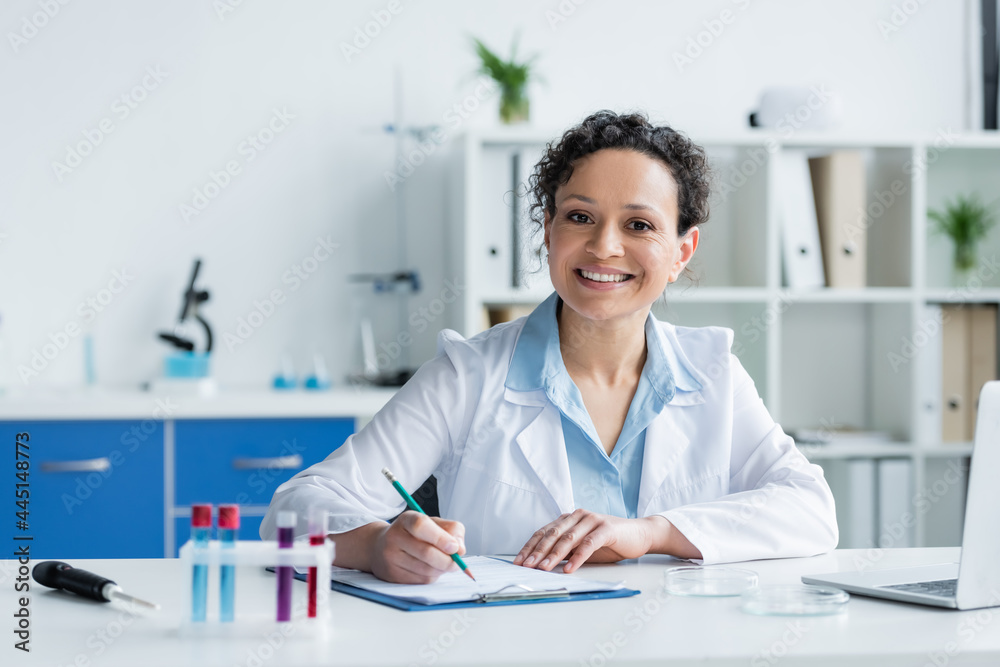 Smiling african american scientist writing on clipboard near blurred test tubes and laptop Smiling african american scientist writing on clipboard near blurred test tubes and laptop