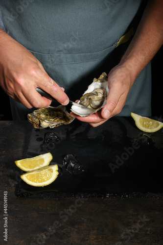 A man s hands are shucking fresh oysters with a blade of knife  Opening the hollow and flat oysters. Chef opens raw oysters in the restaurant  lifestyle. ready to eat on a dark background  luxury food