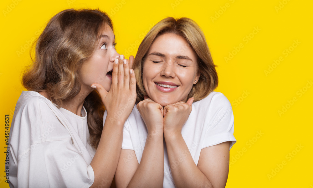 Blonde woman is whispering to her friend something while posing on a yellow studio wall Blonde woman is whispering to her friend something while posing on a yellow studio wall