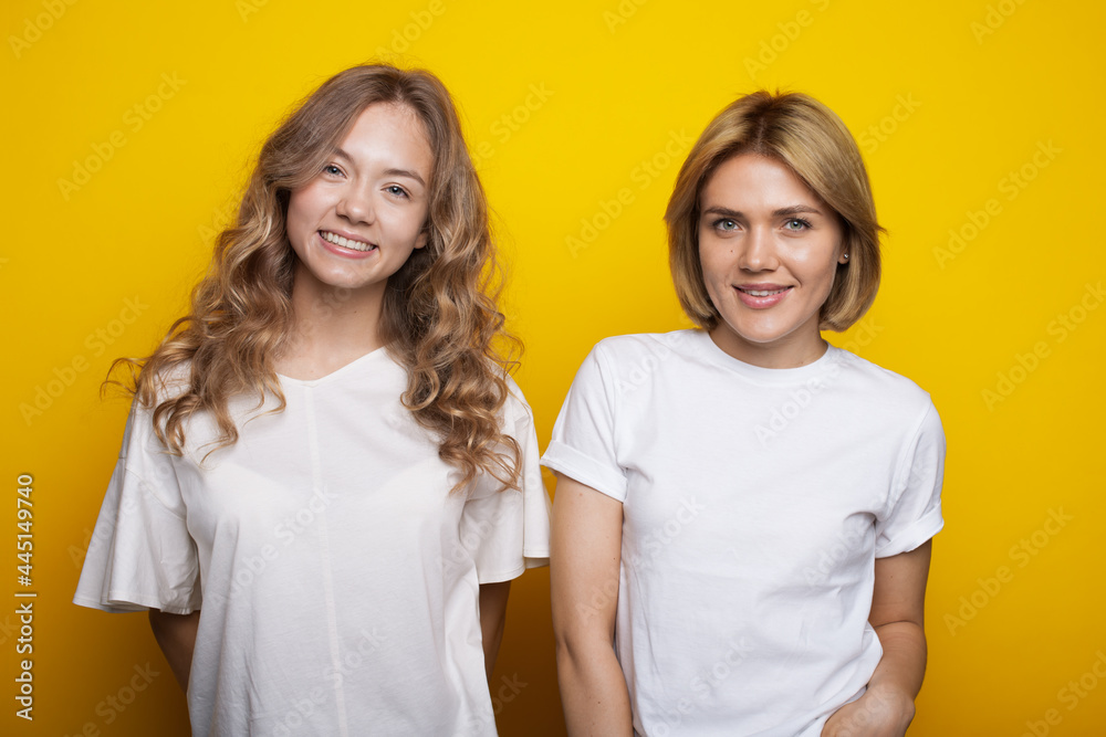 blonde woman and her curly sister are smiling at camera on a yellow studio wall in the same clothes blonde woman and her curly sister are smiling at camera on a yellow studio wall in the same clothes
