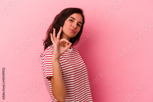 Young caucasian woman isolated on pink background winks an eye and holds an okay gesture with hand.
