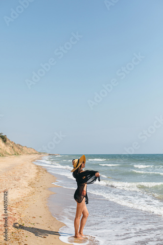 Beautiful carefree woman standing on sandy beach at sea waves and relaxing. Summer vacation. Stylish young female in light black shirt and straw hat enjoying tropical vacation. Space for text