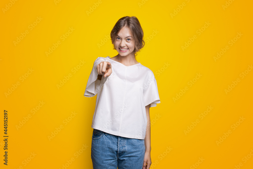 Smiling blonde lady is pointing at camera smiling on a yellow studio wall in a t-shirt