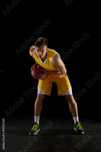 Portrait of young man, basketball player with a ball isolated on dark black studio background. Advertising concept. Fit Caucasian athlete practicing with ball.