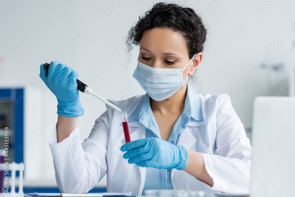 African american scientist in medical mask and latex gloves holding electronic pipette and test tube near blurred laptop African american scientist in medical mask and latex gloves holding electronic pipette and test tube near blurred laptop