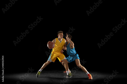 Two basketball players in action and motion isolated on dark black studio background. Advertising concept. Strong Caucasian athletes practicing with basketball ball.