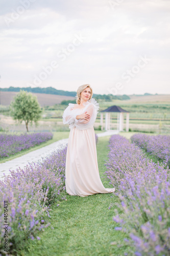 Beautiful middle aged blond Caucasian woman in a field of lavender. Woman in long elegant white dress walks on the lavender field.