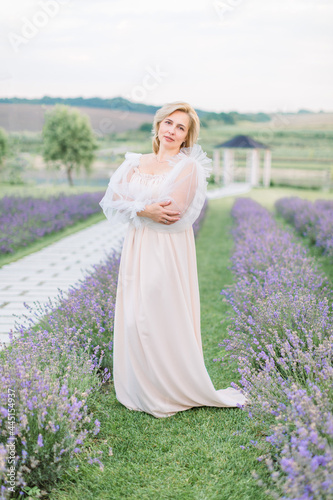 Stunning mature blond lady wearing white long fashionable dress, walking in spring or summer lavender field