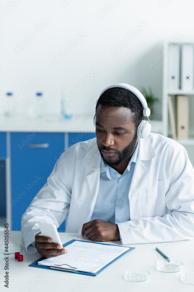 African american scientist in headphones using smartphone near petri dishes and clipboard African american scientist in headphones using smartphone near petri dishes and clipboard