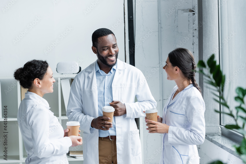 Obraz premium Cheerful interracial doctors holding paper cups and talking in hospital