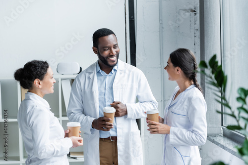 Cheerful interracial doctors holding paper cups and talking in hospital
